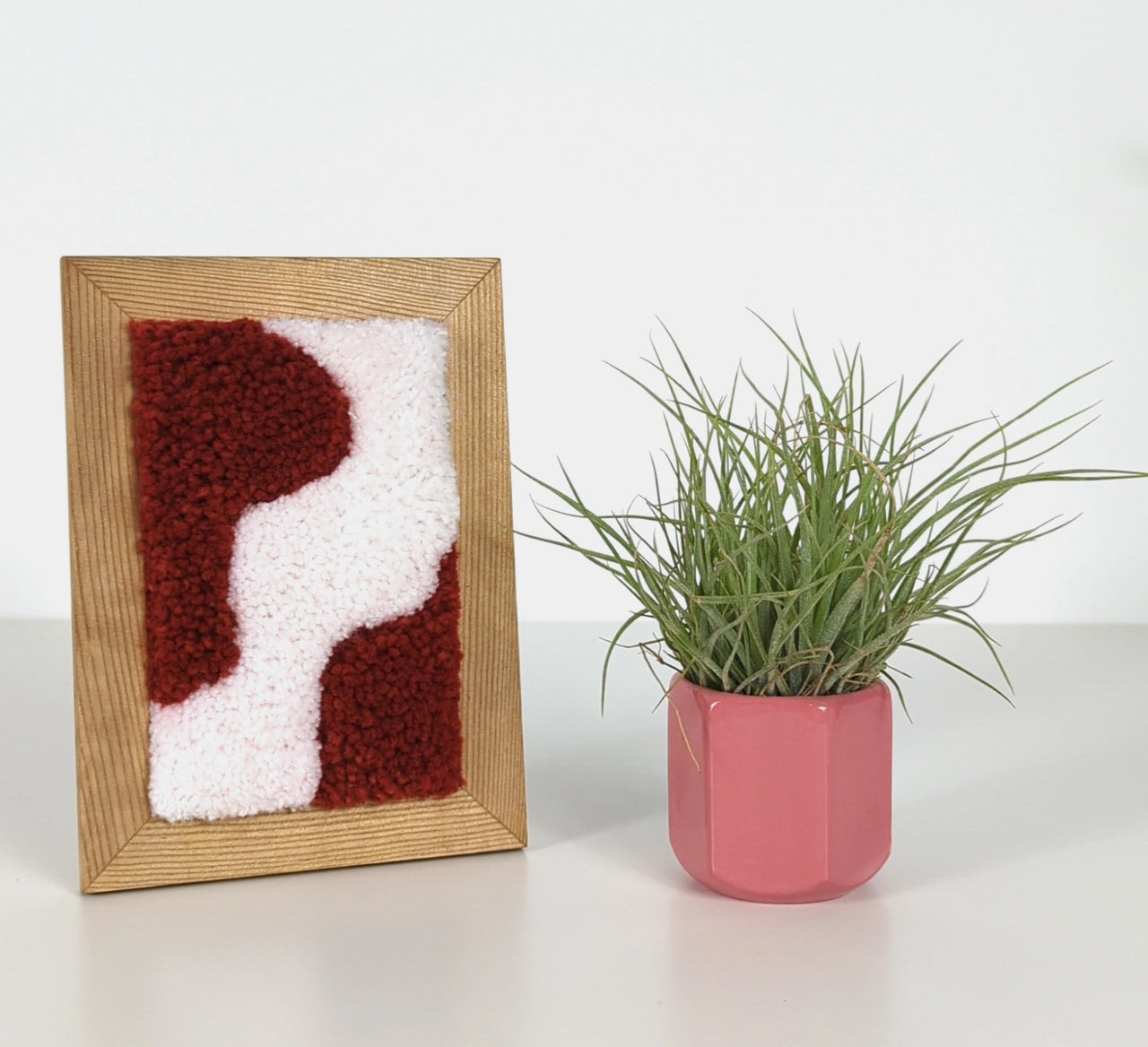 Tableau tufté format A6 blanc et rouge posé à côté d'un petit pot rose contenant une plante verte.